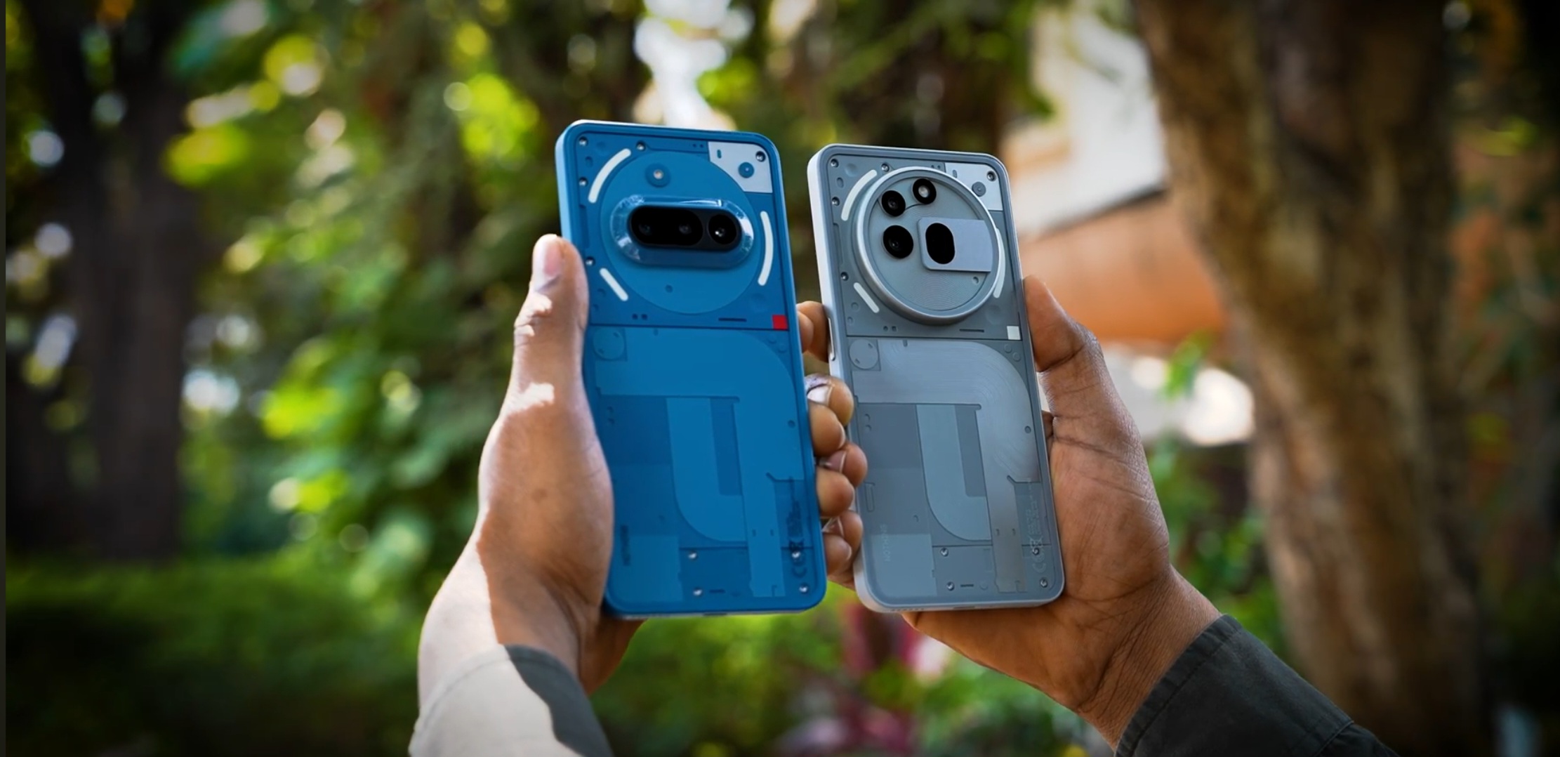 A person holding two smartphones outdoors—Nothing Phone (3a) in blue and Realme 3a Pro+ in silver—both featuring unique transparent back panel designs with visible internal elements.